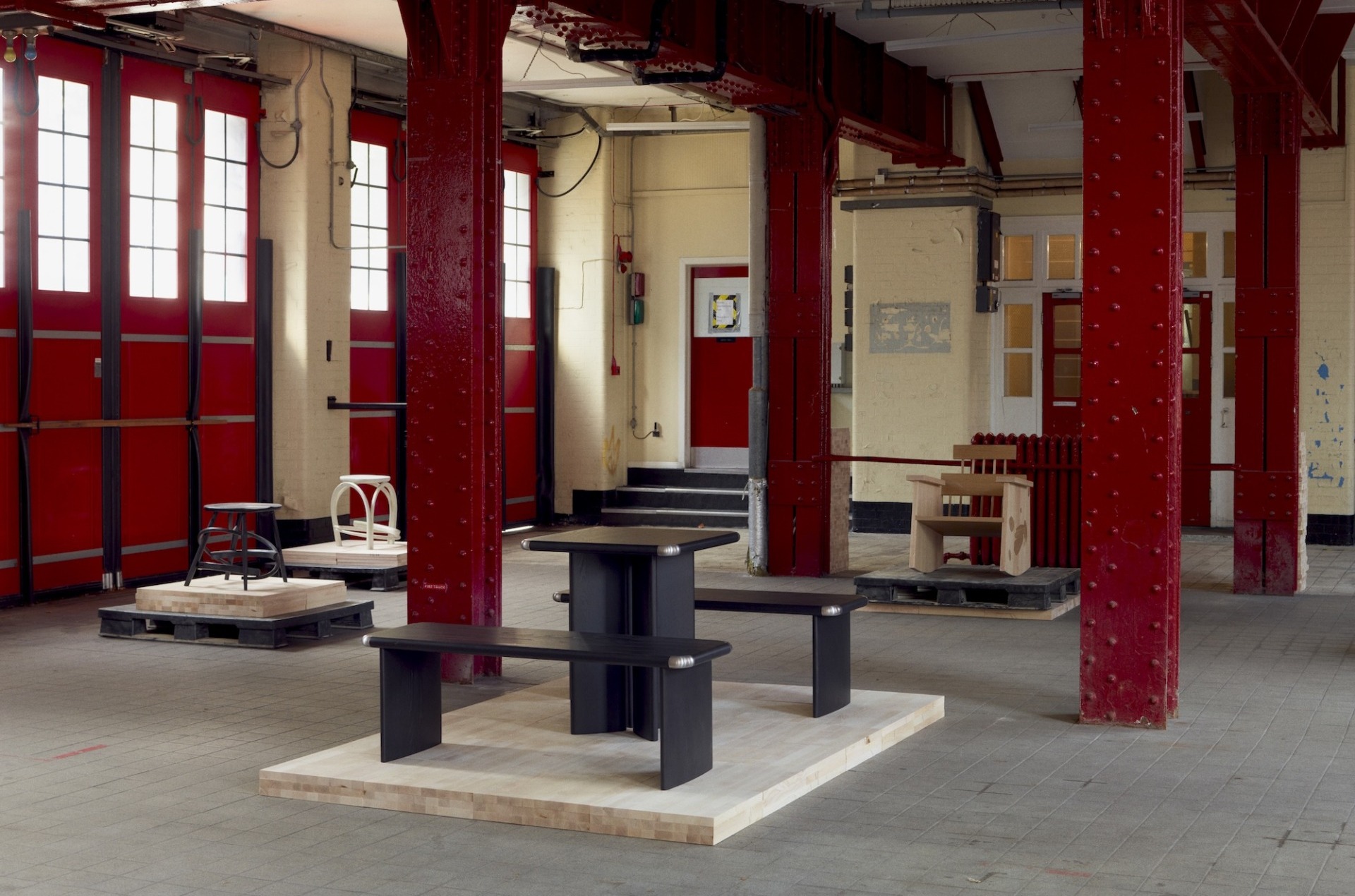 Industrial interior with red beams, black tables on wooden platforms, wide windows, and scattered chairs in a spacious room.
