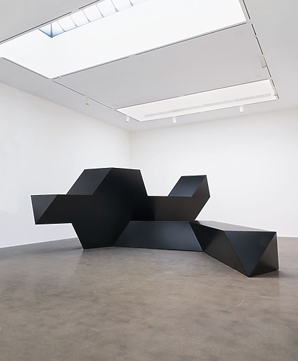 Geometric black sculpture displayed in a modern white gallery space with skylights.