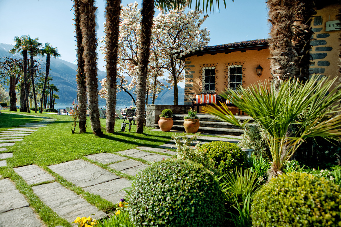 Lakeside garden with palm trees, stone pathway, and quaint house overlooking mountains on a sunny day.