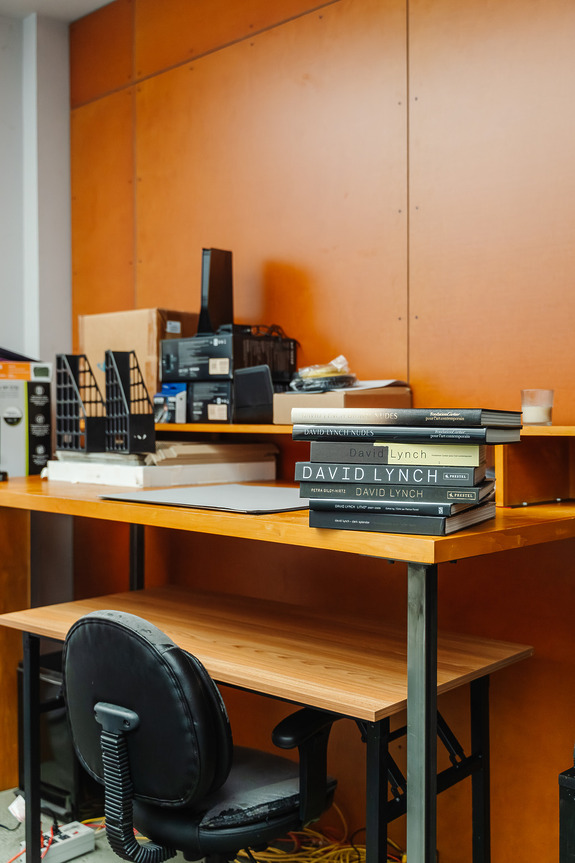 A cluttered office desk with books, electronic equipment, and a black chair against a wooden wall backdrop.