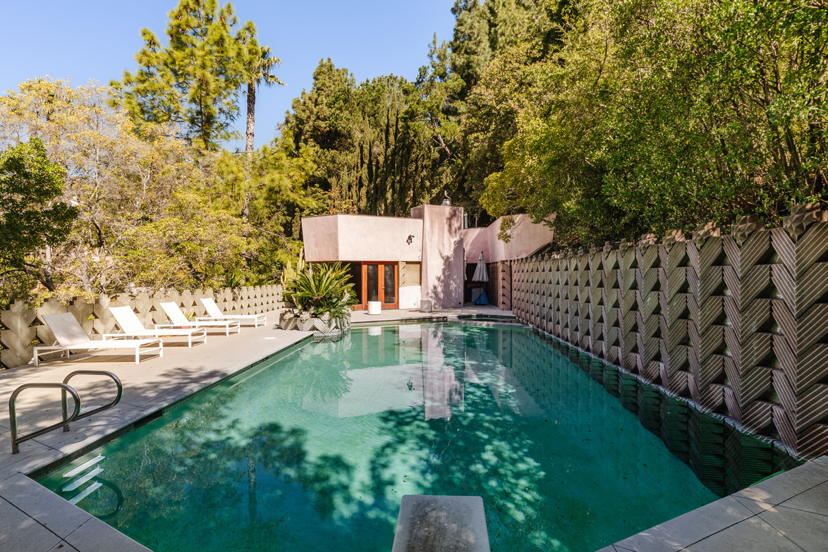 Modern house with a uniquely textured wall, surrounded by lush trees, featuring a serene pool and lounge chairs.