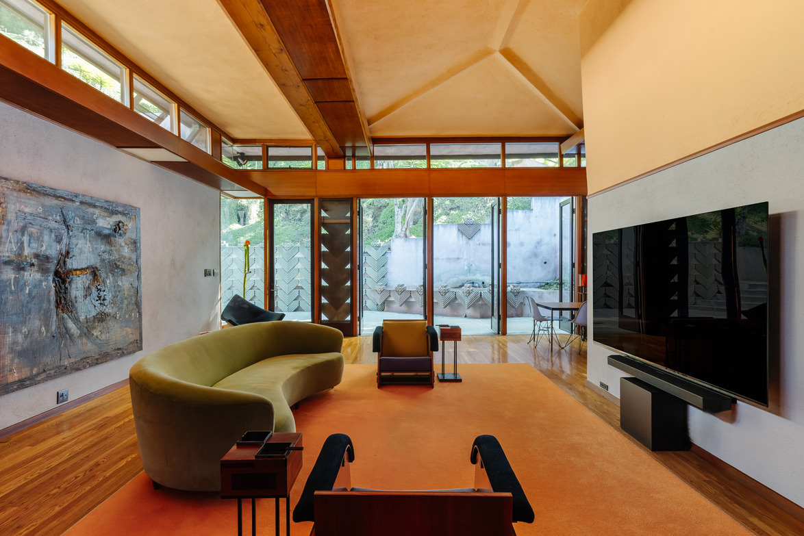 Mid-century modern living room with curved green sofa, large windows, orange carpet, and abstract wall art.