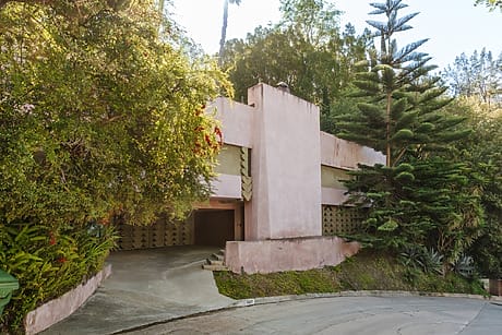 Pink stucco house with modern design in lush, wooded area; driveway and unique geometric wall patterns visible.