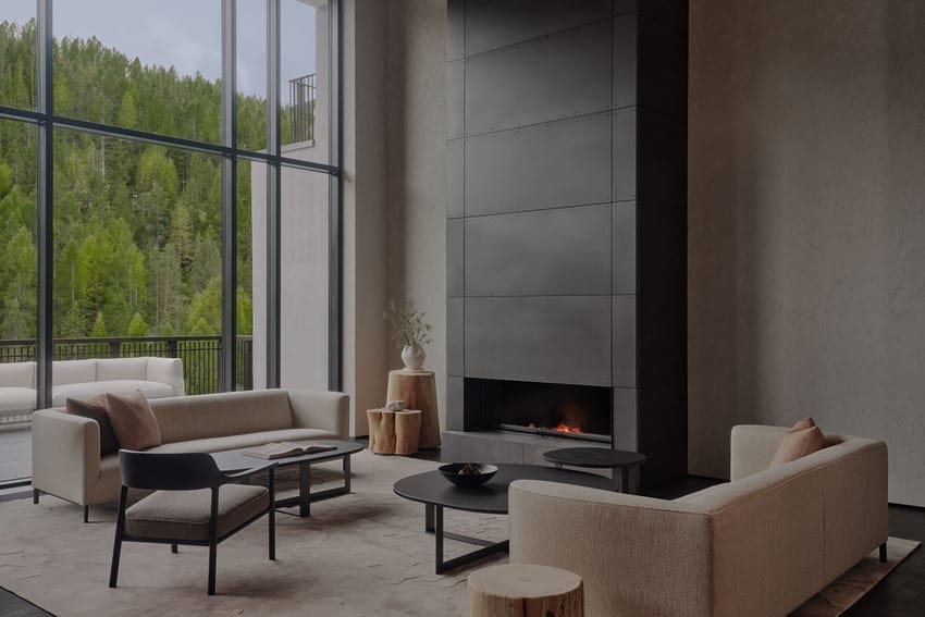 Modern living room with large windows, sleek fireplace, neutral sofas, coffee tables, and chairs, overlooking green forest.
