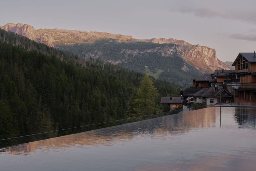 Infinity pool overlooking forested mountains and rustic buildings at sunset.