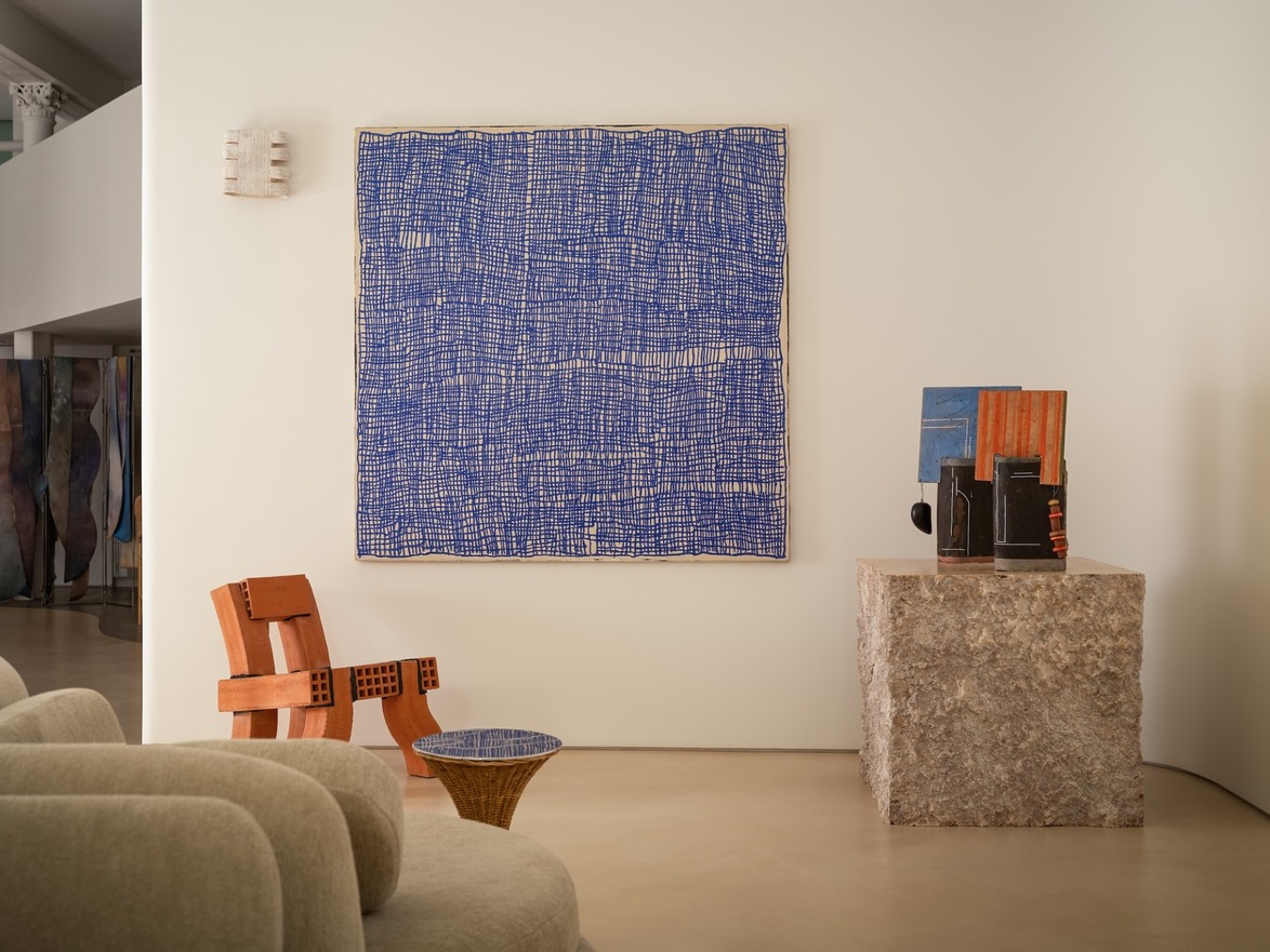 Contemporary art gallery interior with blue abstract painting, modern wooden chair, and sculptures on stone pedestal.