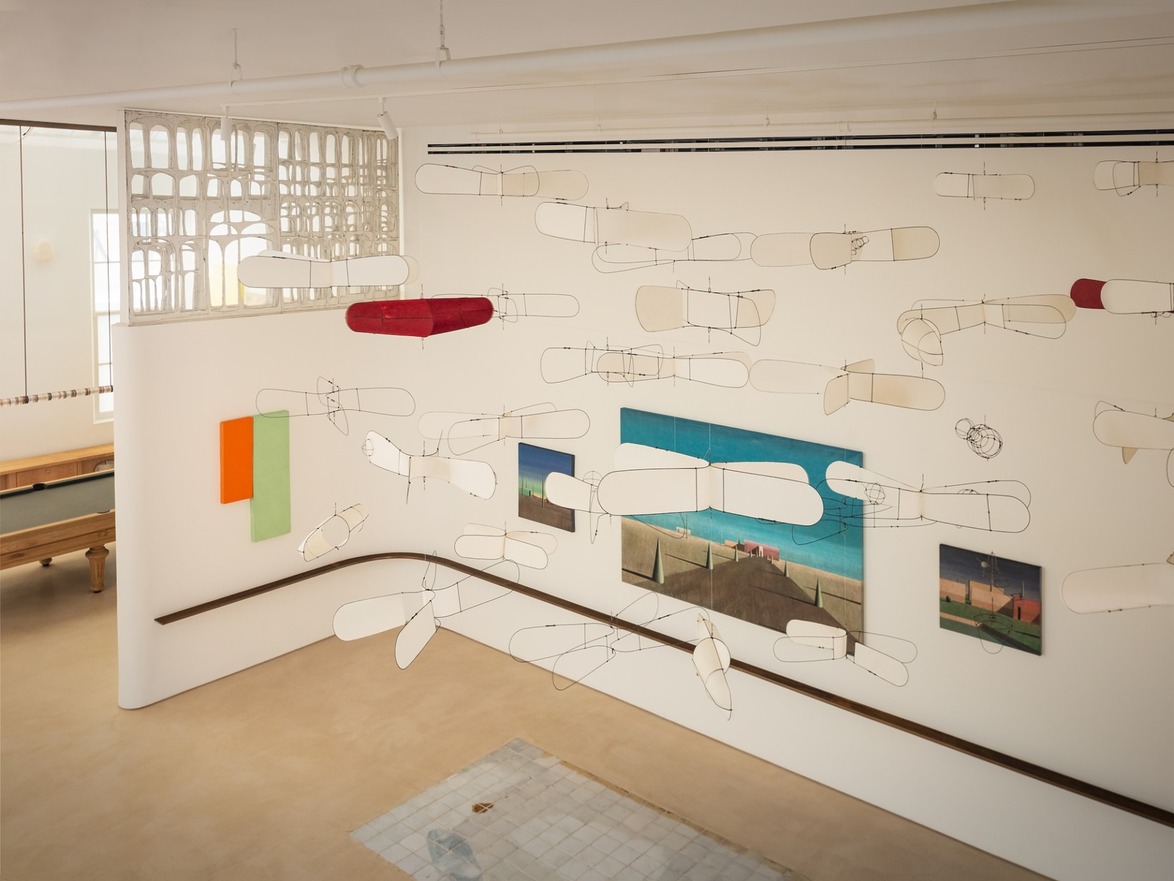 Art installation featuring abstract mobile sculptures and colorful paintings in a modern gallery space with high ceilings.