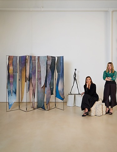 Two women in a gallery with abstract art pieces and a sculpture in the background.