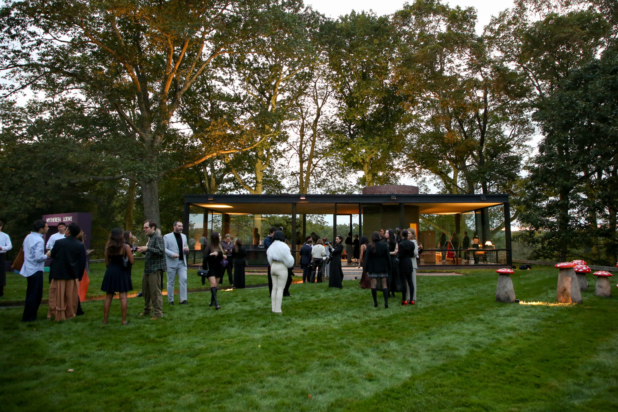 Outdoor gathering at a modern glass house surrounded by trees and grass, with people socializing in formal attire.