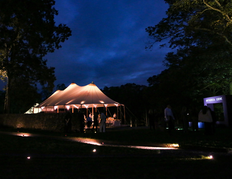 Outdoor evening event with a large illuminated tent surrounded by trees and people gathered nearby under a dark blue sky.