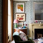 Cozy living room with green armchair, floral wallpaper, framed art, and ornate fireplace mantel.
