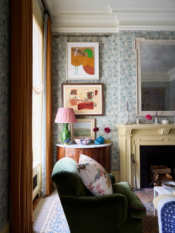 Cozy living room with green armchair, floral wallpaper, framed art, and ornate fireplace mantel.