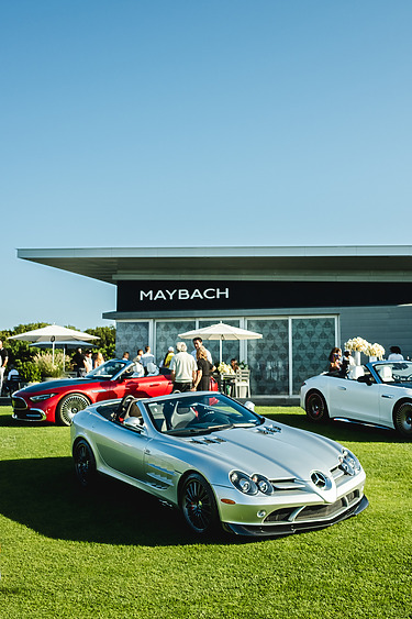 Silver convertible sports car on display at an outdoor car event with people around and Maybach building in background.