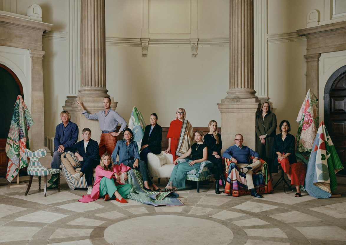 A group of people in colorful clothing sitting and standing in a grand room with columns and quilted textiles.
