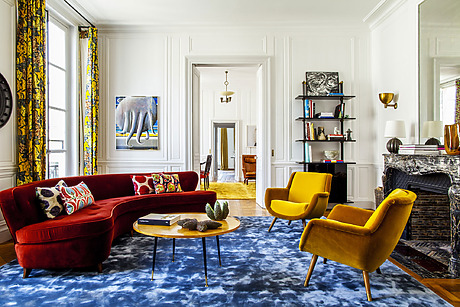 Eclectic living room with red sofa, yellow chairs, blue rug, decorative pillows, and art on white walls.