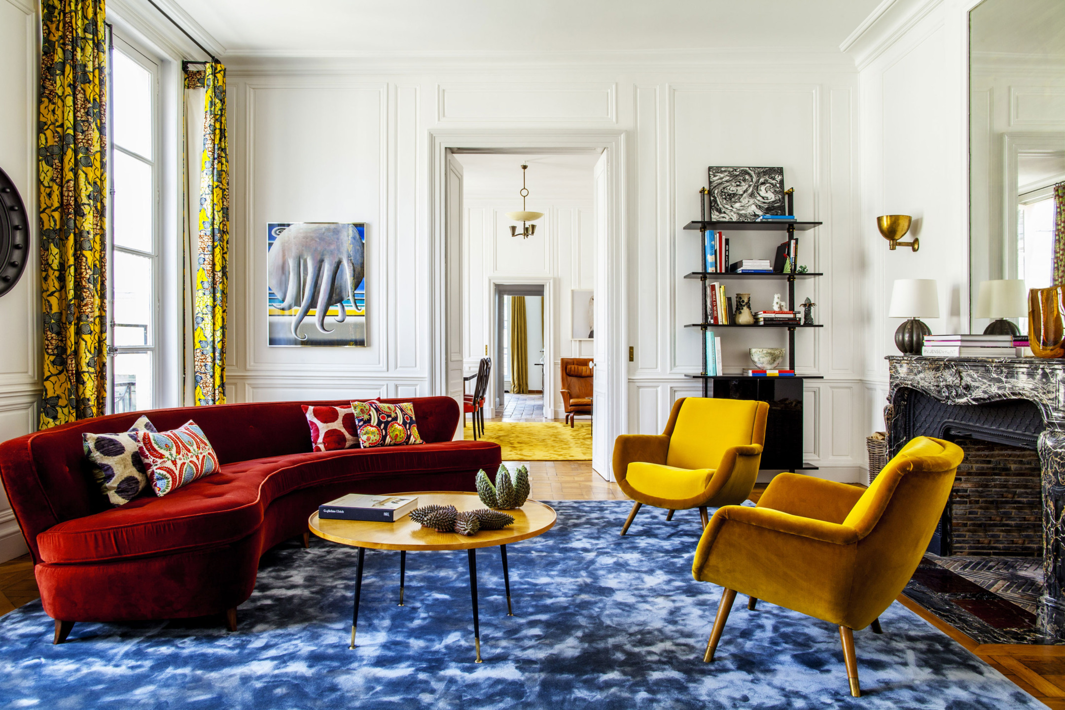 Eclectic living room with red sofa, yellow chairs, blue rug, decorative pillows, and art on white walls.
