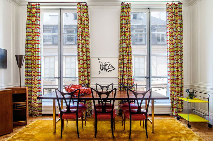 Dining room with colorful curtains, red chairs, yellow rug, and modern art on the wall in a bright space with large windows.