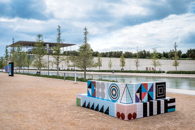 Colorful geometric tiled bench in a modern outdoor park with trees and a building in the background.