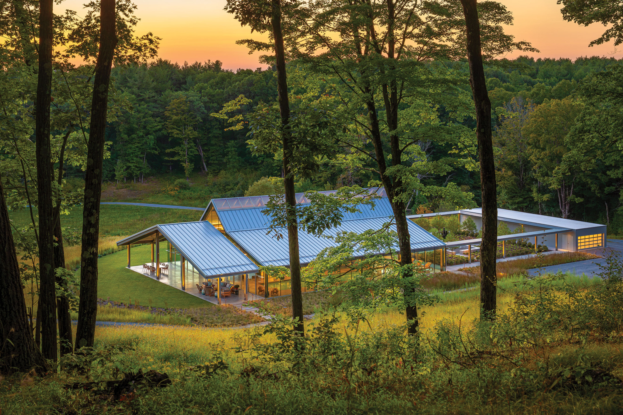 Modern house with a blue roof surrounded by trees at sunset, featuring large glass windows and a green landscape.
