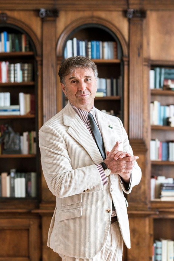 Brunello Cucinelli in a striped suit standing in front of a wooden bookshelf filled with books.