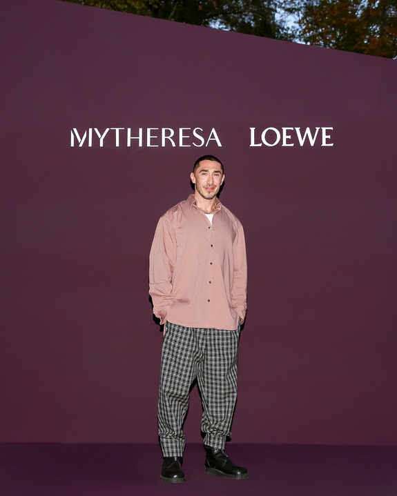 Person standing in front of a purple background with "Mytheresa Loewe" text, wearing a pink shirt and checkered pants.