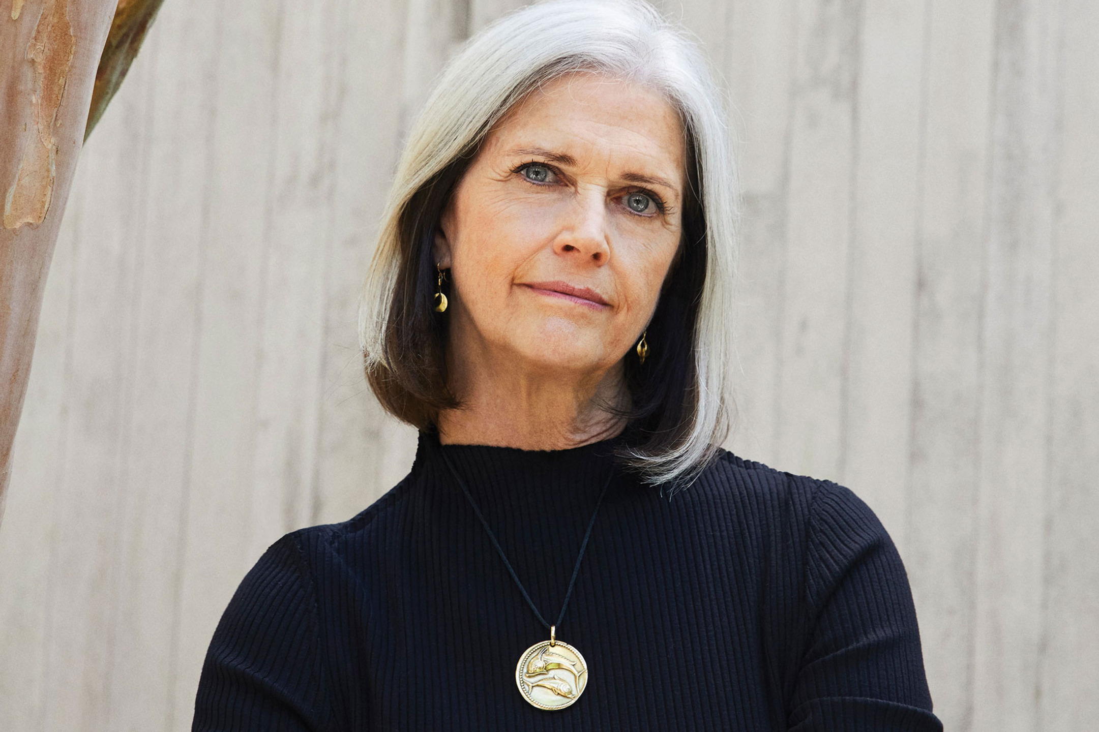 Woman with gray hair wearing black sweater and gold necklace against a wooden background