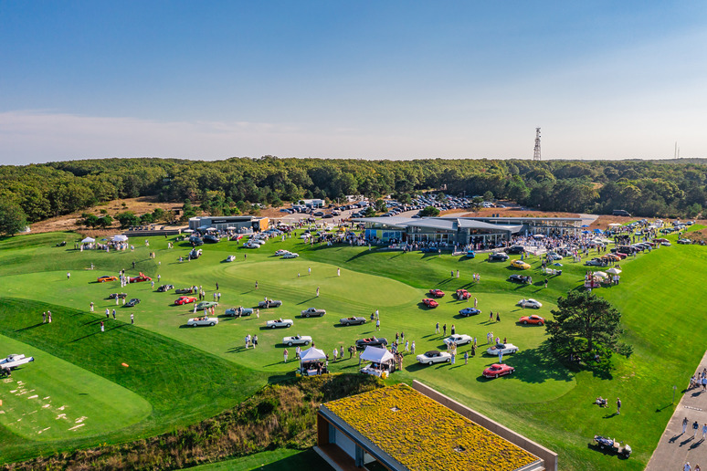 Aerial view of a golf course with people gathering at an outdoor event, showcasing classic cars and green lawns.