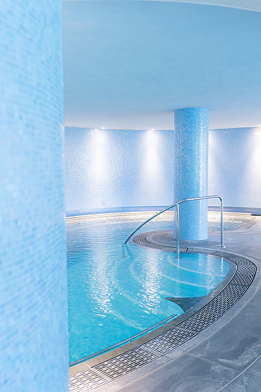 Indoor circular swimming pool with blue mosaic tiles and metal handrail in a bright, softly lit room.