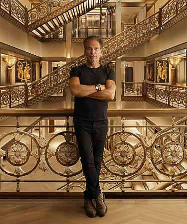 Man standing with arms crossed in ornate building featuring intricate metalwork and multiple staircases in the background.