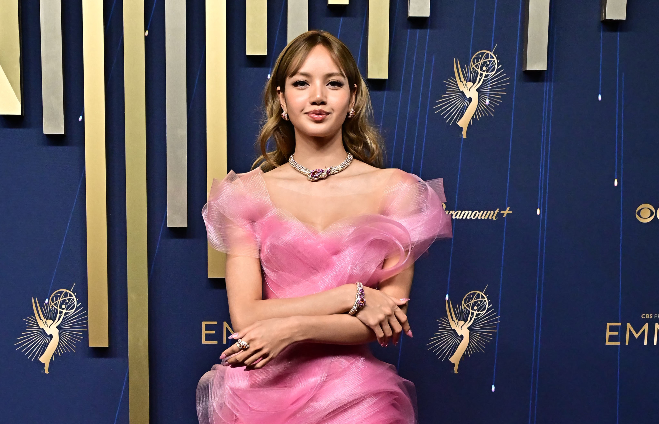 Person in a pink dress standing with arms crossed at a formal event with decorative backdrop.