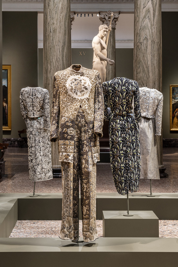 Exhibit of intricately patterned outfits displayed on stands in an ornate gallery setting with classical sculptures.