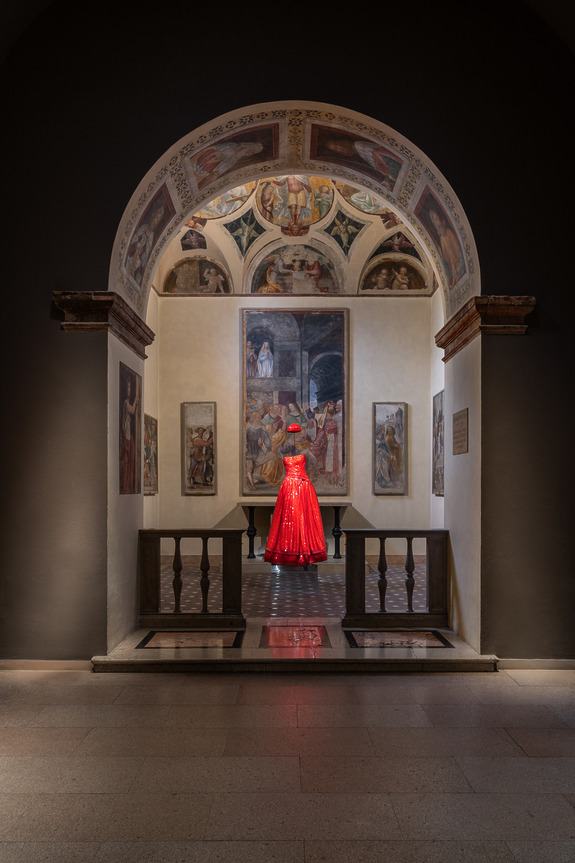 A vivid red gown displayed under an ornate arched ceiling with classical paintings in a dimly lit gallery room.