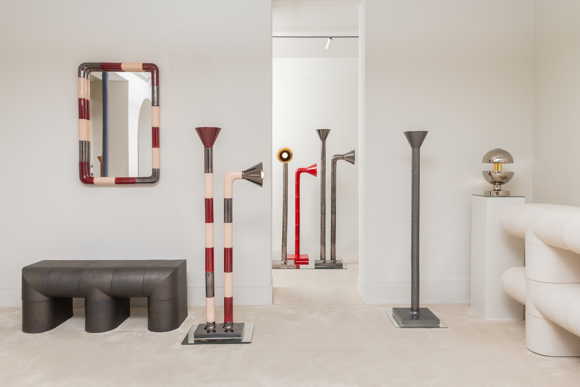 Modern art gallery with colorful geometric sculptures, a striped mirror, and a unique lamp on a minimalist white wall.