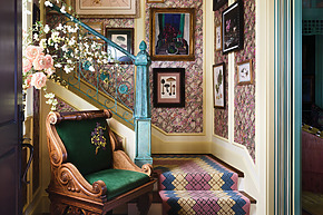Eclectic staircase with patterned wallpaper, colorful carpet, framed art, and an ornate wooden chair with floral embroidery.
