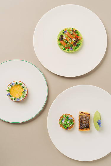 Three plates of gourmet dishes including plated fish, garnished salads, and a green sauce on a beige surface.