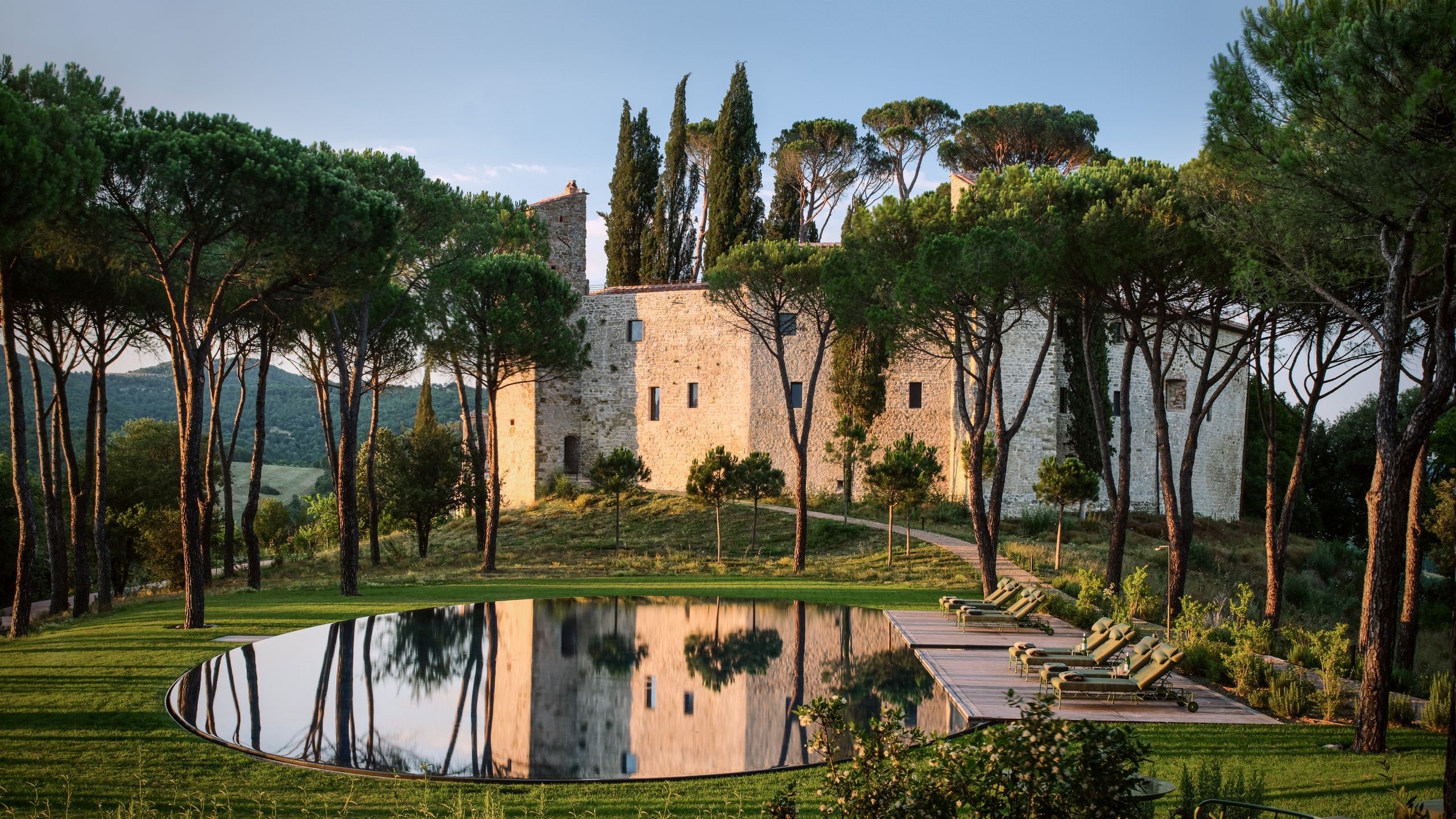 Historic stone villa surrounded by trees with a circular pool reflecting the landscape at sunset.