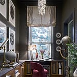 Elegant study with chandelier, desk by window, plush chair, globe decor, bookshelves, and framed art on dark textured walls.