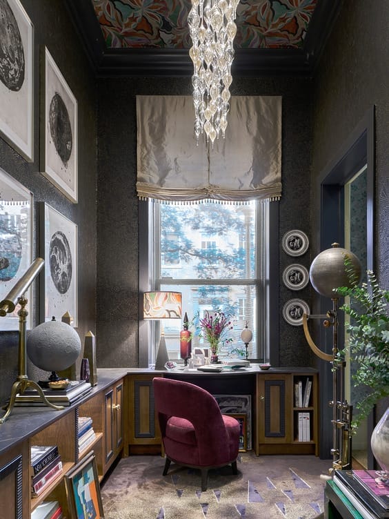 Elegant study with chandelier, desk by window, plush chair, globe decor, bookshelves, and framed art on dark textured walls.