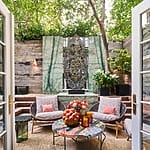 Cozy outdoor patio with colorful table, wicker chairs, and lush green plants surrounded by rustic wood and stone walls.