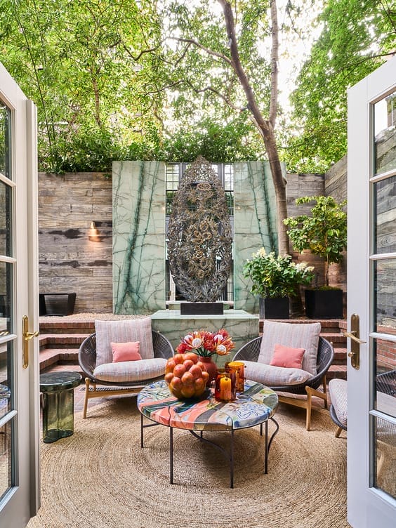 Cozy outdoor patio with colorful table, wicker chairs, and lush green plants surrounded by rustic wood and stone walls.