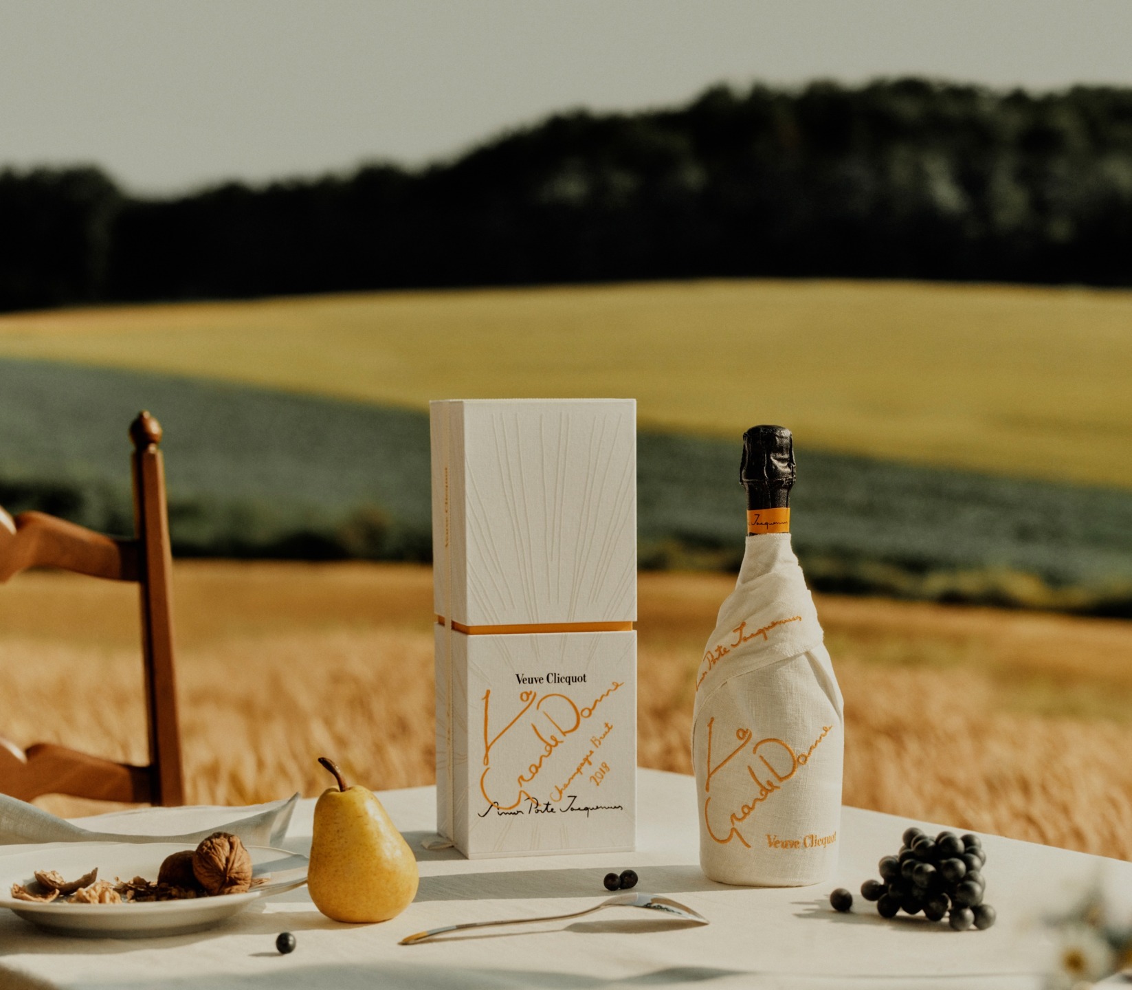 Champagne bottle with white label, box, pear, nuts, and grapes on a table in a field with greenery in the background.