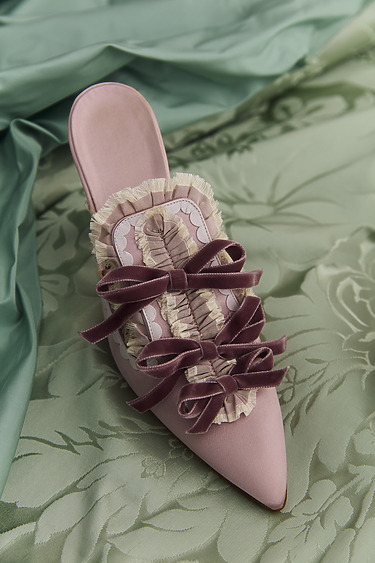 Pink pointed-toe shoe with ruffled details and purple bows on a green floral fabric background.