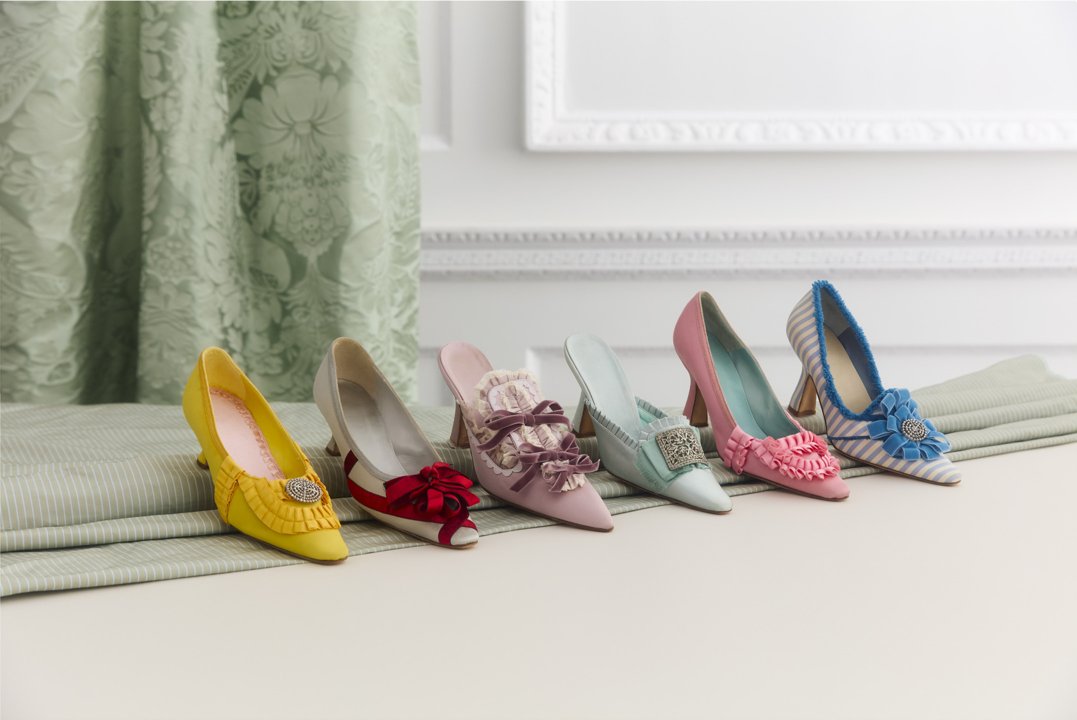 Colorful high-heeled shoes with bows and embellishments displayed on a striped fabric in front of floral curtains.