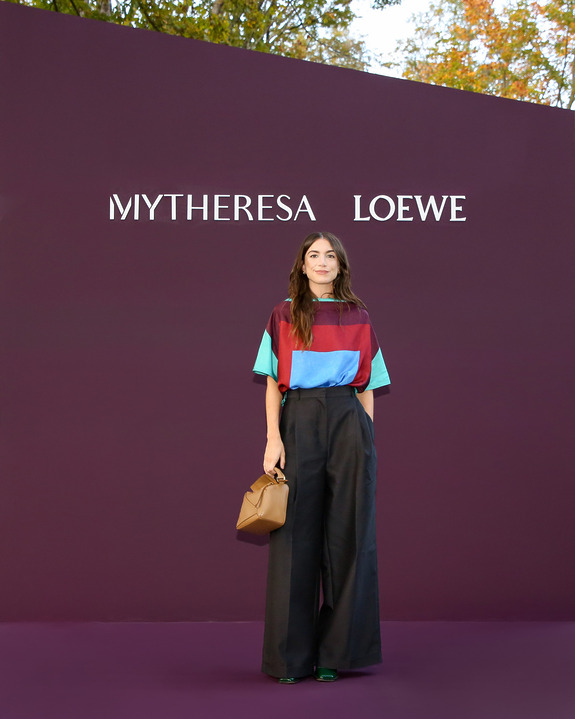Person in colorful shirt and wide-leg pants stands in front of a maroon Mytheresa Loewe backdrop, holding a brown bag.