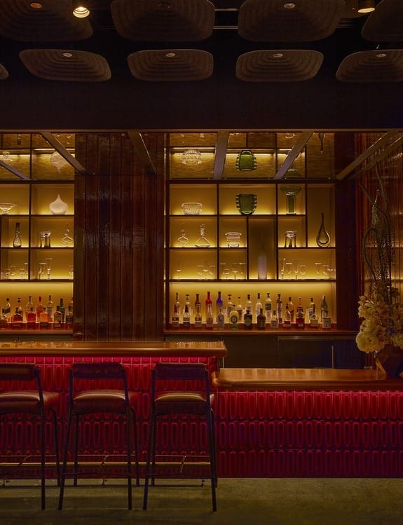Dimly lit bar with red counter, three stools, and shelves of bottles and glassware in the background.