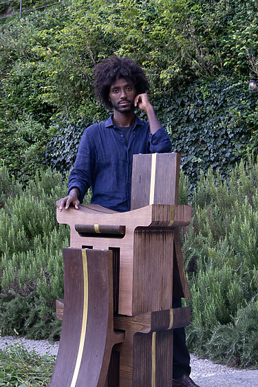 Person standing beside a uniquely designed wooden sculpture in a garden setting with lush greenery in the background.