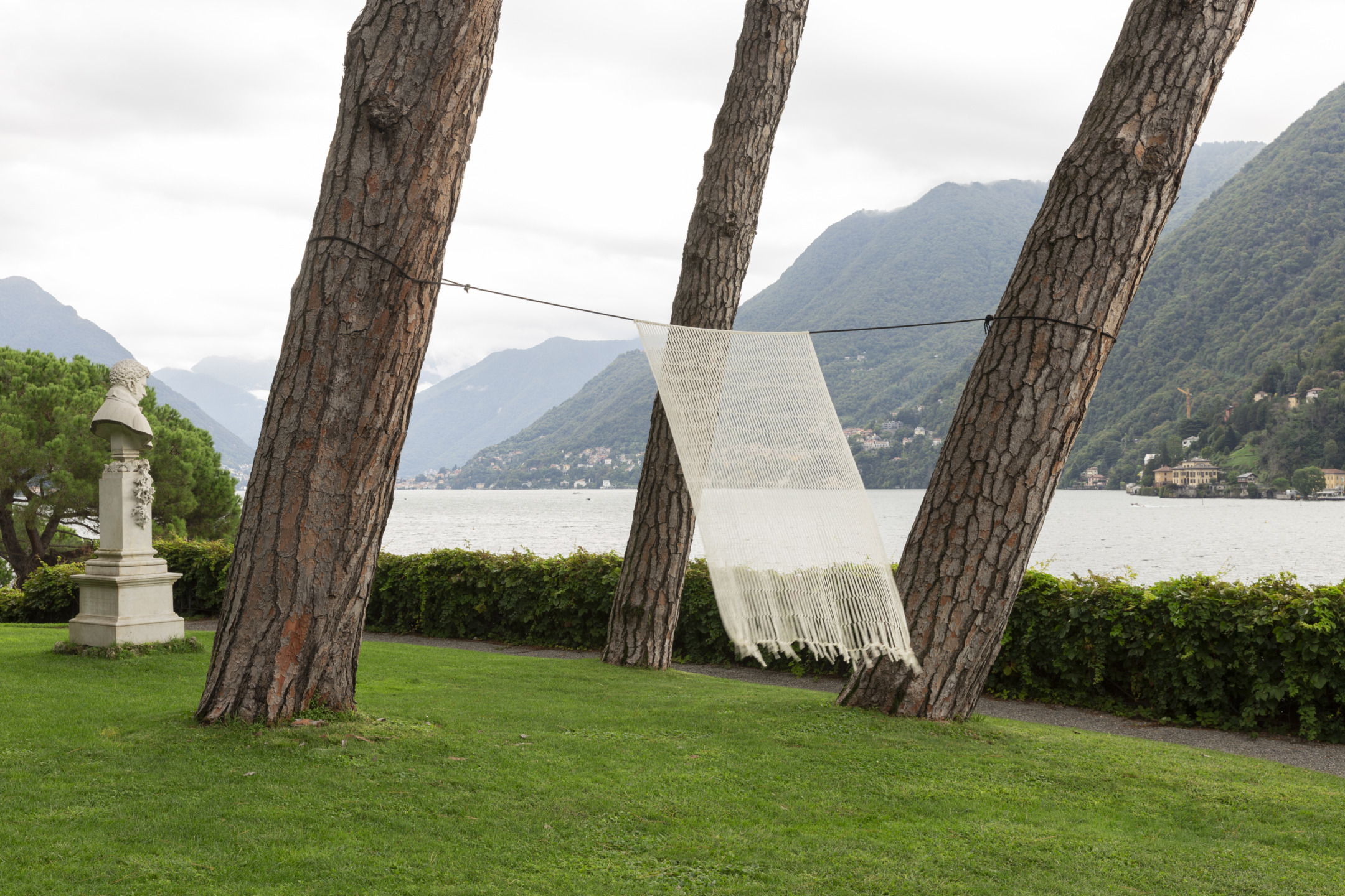 Art installation with a woven textile hanging between trees by a lake with mountains in the background.