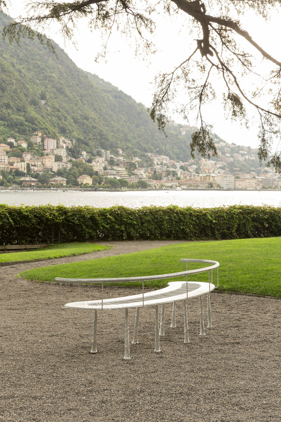 Curved bench on a gravel path by a lake, lush green grass, mountains with houses in the background, overhanging tree branches.