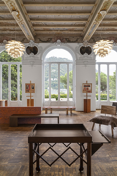 Spacious room with ornate ceiling, large arched windows with a scenic view, and art displayed on easels with wooden furniture.