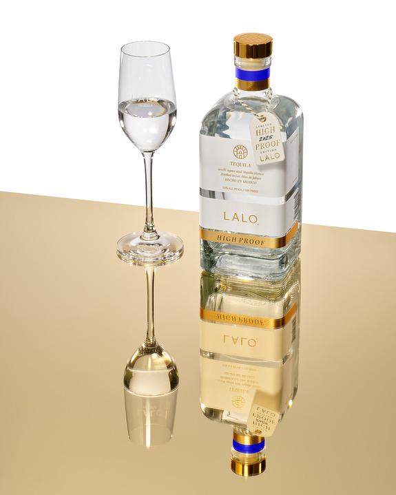 Bottle of Lalo high-proof tequila on reflective surface with filled glass next to it